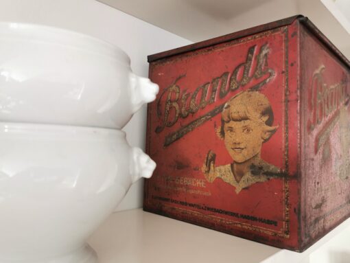 Alte Werbevitrine von Brandt mit unverkennbarem Markenschriftzug und Mädchen, das Gebäck in der Hand hält. Dose steht in Regal neben zwei Löwenkopfterrinen
