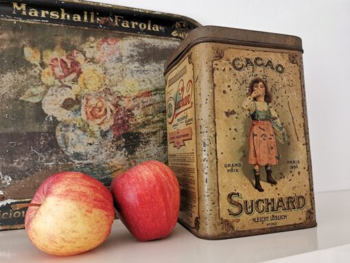 Alte Blechdose Suchard Cacao, dekoriert mit zwei Äpfeln und einem antiken Metall-Tablett, das wie die Dose lithografiert ist.
