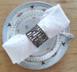Antiker silber Serviettenring, durchbrochen mit Rosendekor und Silberperlen. Der Ring ist auf einer Serviette aufgezogen die quer über einer Teetasse liegt, darunter eine Untertasse und ein Teller, jeweils mit Blumen- bzw. Rosenmuster. Das Foto wurde frontal von oben aufgenommen.