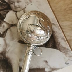 Nahaufnahme Babyrassel aus Silber mit Schwanenmotiv: Schwann auf einem Seerosenteich. Ein Teil des Stiels ist ebenfalls zu sehen siowie ein Bildausschnitt des antiken Kinderfotos, auf dem die Rassel dekoriert wurde.