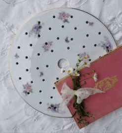 Weißer alter Porzellan Untersetzer mit Streublumendekor: Rosa und lila Röschchen. Die Oberfläche des Untersetzers hat Abtropflöcher. In der MItte ist ein großes Loch