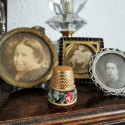 Alter Fingerhut aus Messing mit bezaubernder Petit Point Stickerei steht inmitten von drei Miniatur Standbilderrahmen mit Porträtfotos um die Jahrhundertwende. Im Hintergund ist noch ein Stück eines Kristall-Lampenfußes zu erkennen.