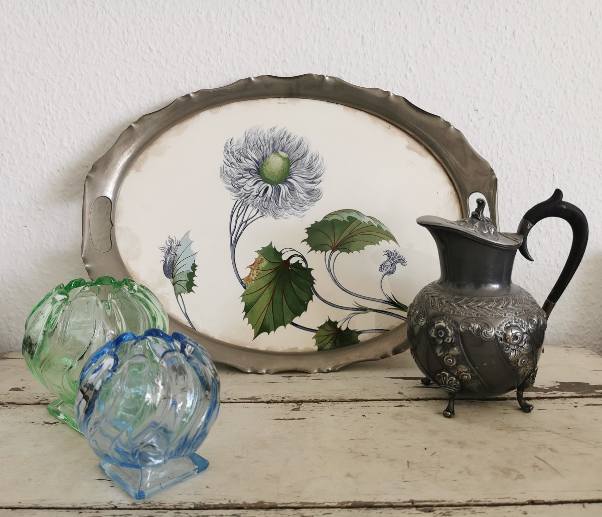 Antikes Jugendstil Tablett_ Keramik mit Metallmontur, mittig eine blaue Blume. Das Tablett lehnt an der Wand. Davor steht rechts eine antike kleine Metall-Kanne mit Rosen auf dem Kannenkorpus und einem schwarzen Griff. Links neben dem Tablett stehen zwei Kugelvasen aus grünem und blauem Klarglas.