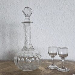 Antike Karaffe für Likör auf einem Podest. Die Karaffe hat eine bauchige Form und einen langen Hals. Neben der Karaffe stehen zwei kleine Likörgläser.