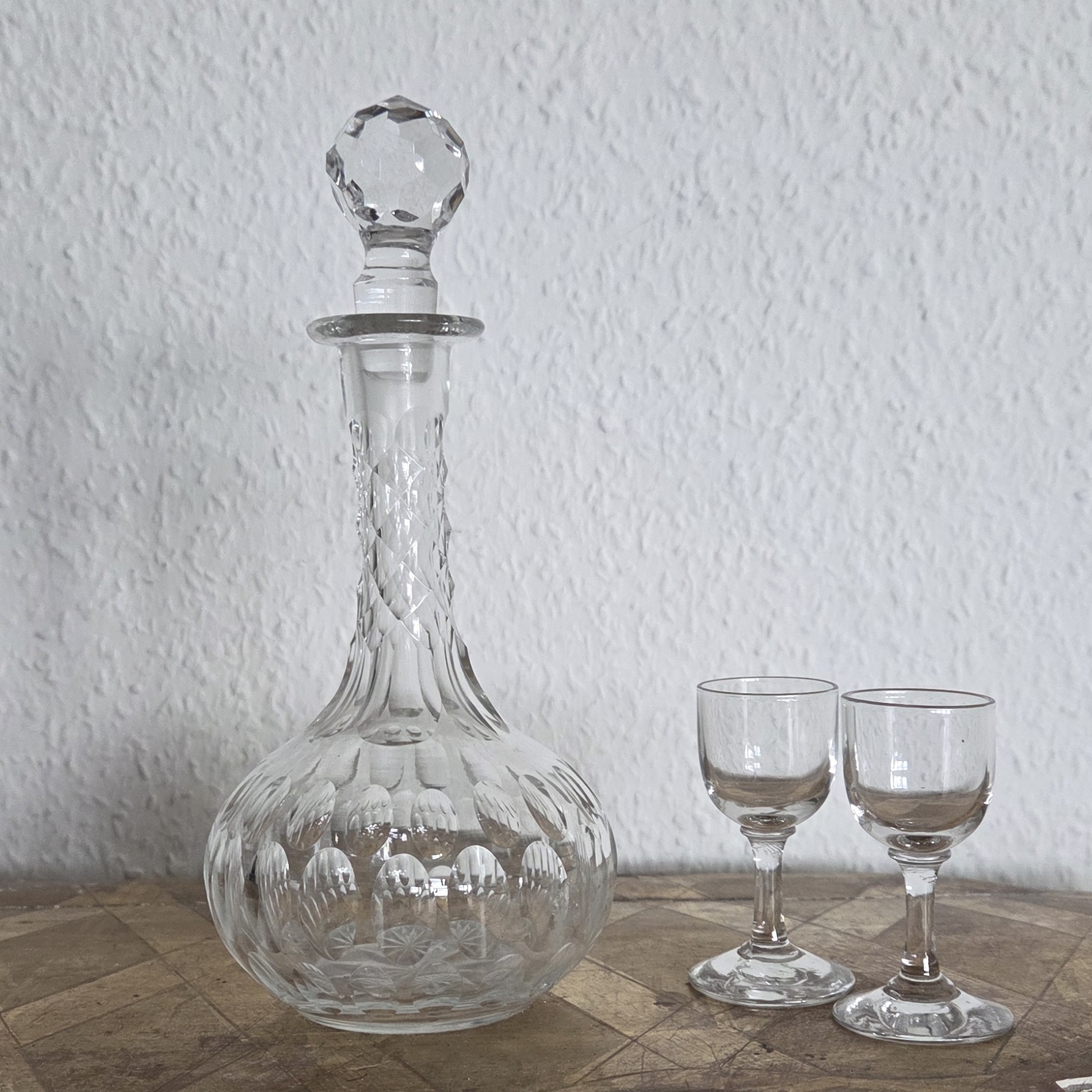 Antike Karaffe für Likör auf einem Podest. Die Karaffe hat eine bauchige Form und einen langen Hals. Neben der Karaffe stehen zwei kleine Likörgläser.