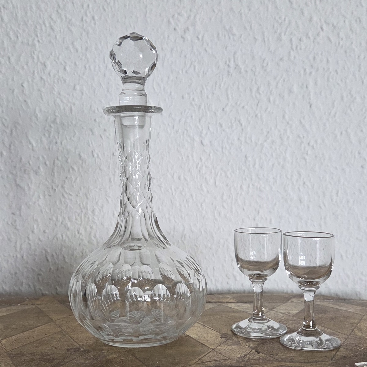 Antike Karaffe für Likör auf einem Podest. Die Karaffe hat eine bauchige Form und einen langen Hals. Neben der Karaffe stehen zwei kleine Likörgläser.